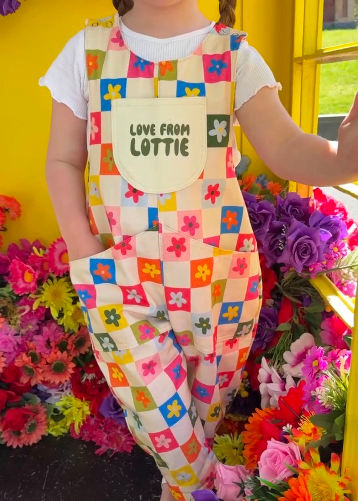 Child wearing a colorful checkered romper with a floral design, standing in front of flowers.