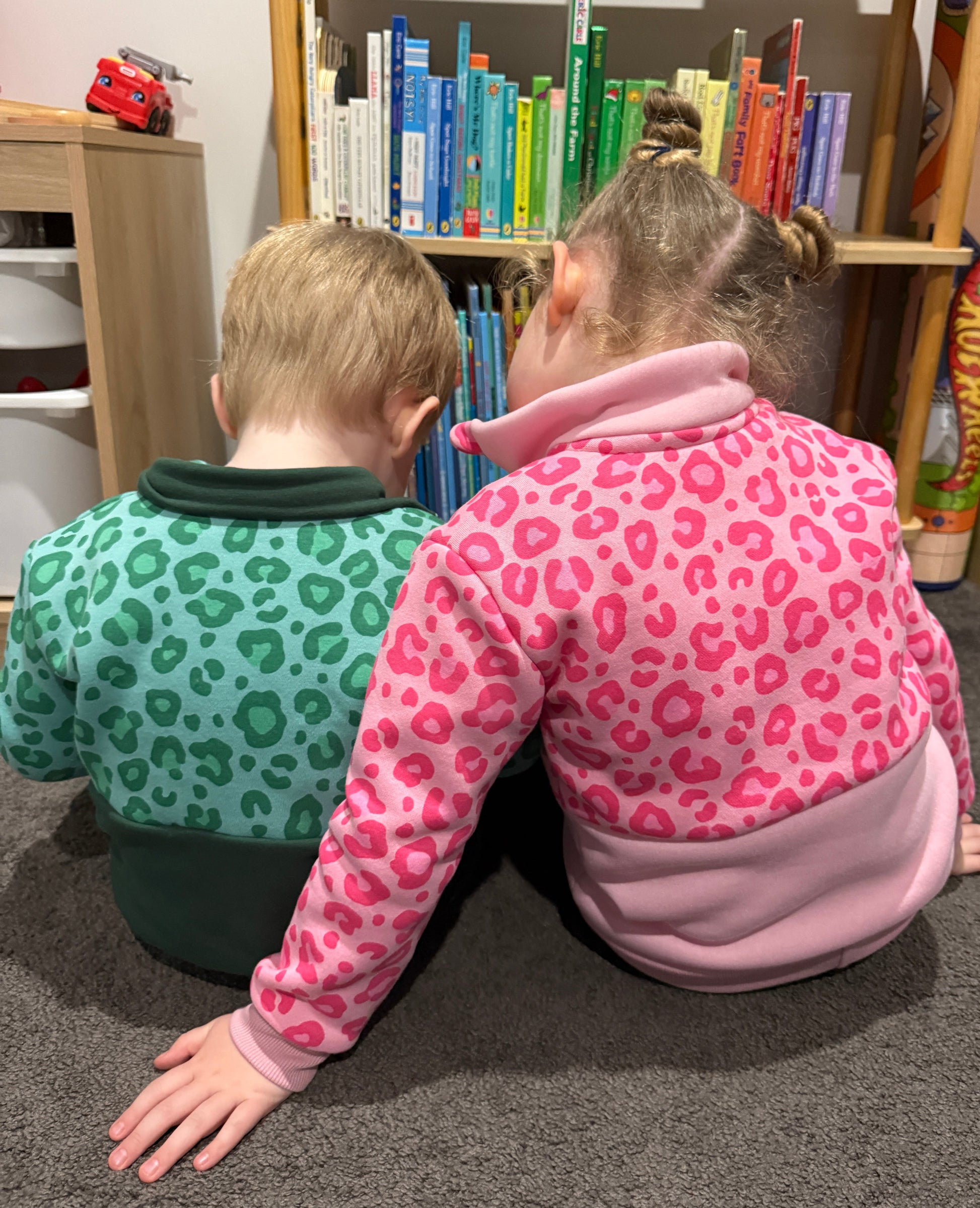 Two children in leopard print jumpers looking at books on a shelf.
