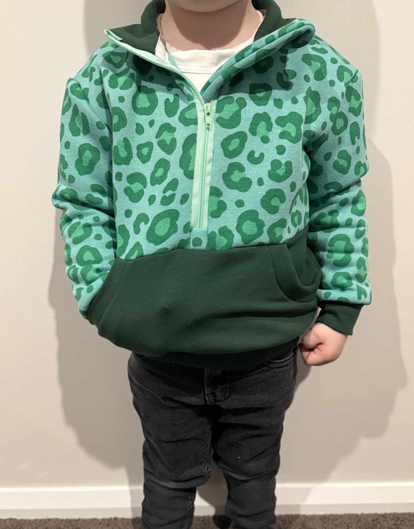 Child wearing a green leopard print jumper and dark pants against a plain wall.
