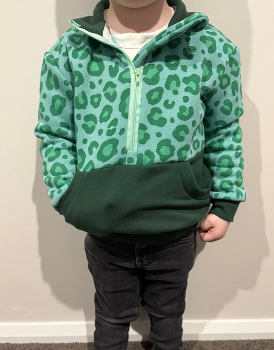 Child wearing a green leopard print jumper and dark pants against a plain wall.
